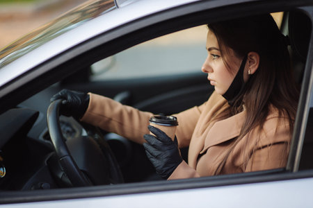 Woman with face mask driving her car with cup of coffee during coronavirus pandemic, Covid-19. Woman pulled of protective mask for drinking cappucinoの写真素材
