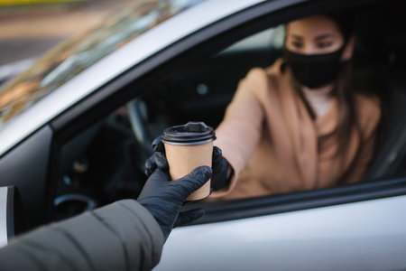 Attractive young woman take cup of coffe while sitting in car. Woman in face mask and gloves. Woman...