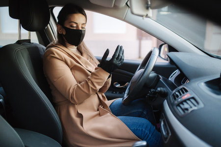 Beautiful young girl in a mask sitting in a car and put on protective gloves. Protective mask against covid-19, driver on a city street during a coronavirus outbreakの写真素材