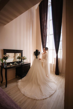 Beautiful young bride in negligee stand by her wedding dress in hotelの写真素材
