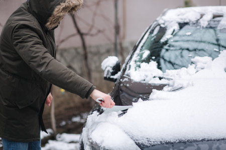 A man is cleaning snowy window on a car with snow scraper. Focus on the scraper. Cold snowy and...