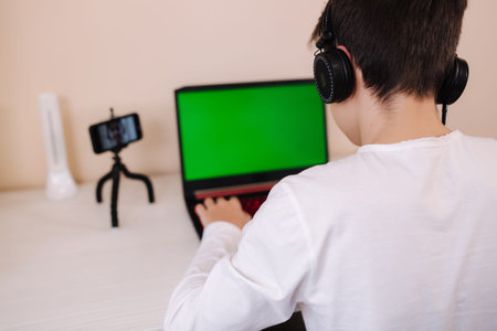 Back view of young boy play gamer on laptop greenscreen. Steamer in earphones capture video on his...