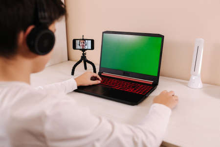 Back view of young boy play game on laptop. Green screen. Steamer in earphones capture video on web camera. Focus of laptopの写真素材