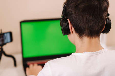Back view of young boy play game on laptop. Green screen. Steamer in earphones capture video on web camera. Close upの写真素材