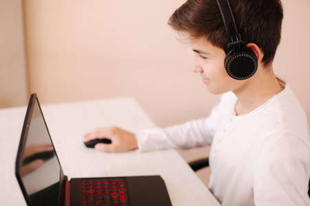 Teenage boy playing game on laptop in his room. Steamer use wireless headphones. Happy gamer play all dayの写真素材