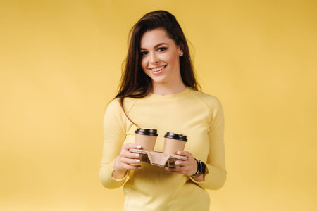 Brunette woman hold two cup of cappuccino on yellow background in studio. Attractive female smiled...
