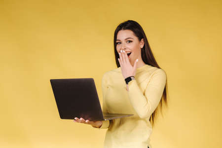Happy surprised woman in yellow sweatshit hold laptop and raised hand to face. Portrait of pretty woman on yellow background.の写真素材