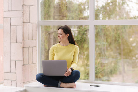 Pretty woman sitting on window sill with laptop and woking. Freelancer at home barefoot. Brunette...