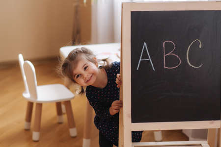 Adorable little girl stand by easel with blackboard and have fun. Cute little kid play and study at home during quarantine. Written ABC on black blackboardの写真素材