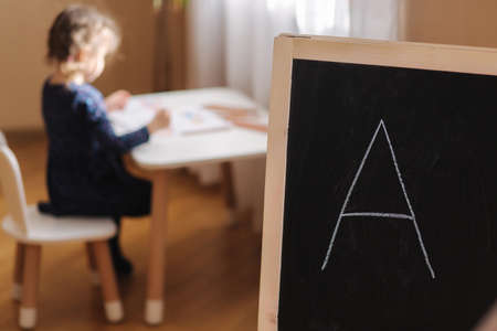 Adorable little girl sitting at the table and play. Cute little kid play and study at home during quarantine. On focus Written letter A on black blackboardの写真素材