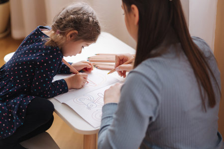 Adirable little girl with her beautiful young mom draws in the album using color pensins. Cute girl paints coloring page at home during quararntine. Happy kid sits at the table by the windowの写真素材