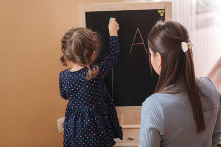 Cute little daughter with her young mom learn writes a letters on the blackboard at home. Side view of female kid in dress learningの写真素材