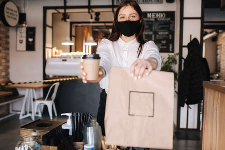 Female cashier with face mask serving coffee to customer, shop open after lockdown. Woman hold packege with food and coffeeの写真素材