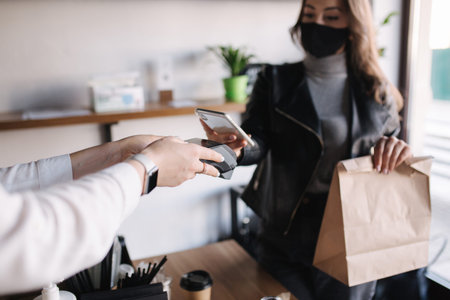 Young woman in mask paying bill through smartphone using NFC technology in a cafe. Female customer...