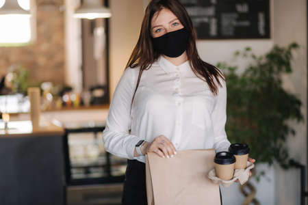 Cashier in face mask holding package with food and two cups of coffee. Contactless payments concept at cafe. Take away food during qurantine.の写真素材