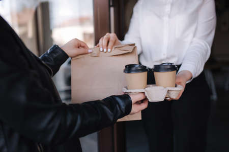 Female cashier in face mask serving coffee and package with food to customer. Coffee shop open after lockdown. Woman hold packege with food and coffee. Focus on handの写真素材