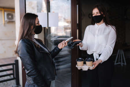 Woman standing outdoors by cafe and paying with smartphone during  pandemic. Cashier hand holding credit card reader machine while client holding phone for NFC payment. Woman take coffeeの写真素材