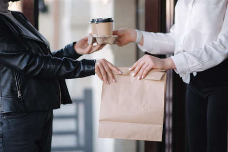 Customer in face mask stand outdoors and take package with coffee and food from cashier in mask at cafe. Take away food. Quarantineの写真素材