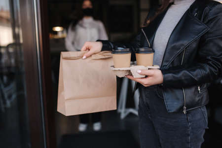 Attractive female customer in face mask standing outdoors by cafe and take away food and coffee. Background of cashier in mask waiting for next customer. New norm during quarantine.の写真素材