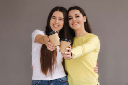 Two attractive girls hold cup of coffee in studio on grey background. Brunette female in yellow sweater. Beautiful females smileの写真素材