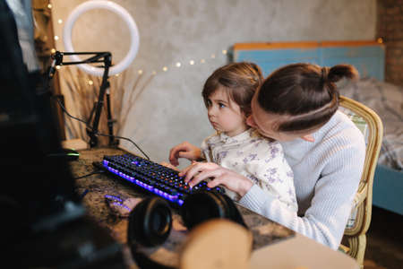 Younger sister sits on brothers knees and put finger on keyboard with neon light. Little girl help brother work on computer. Happy familyの写真素材