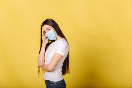 Portrait of young woman in medical mask stand in frond of yellow background. Girl has a headache...