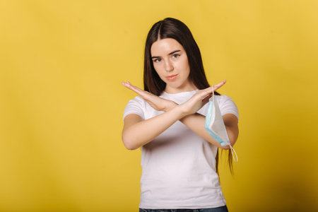 Attractive young woman hold protective mask in hand and think of its necessity. Female cross arms...
