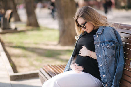 Happy pregnant woman put hands on belly and looking on tummy. Female sitting on the bench in park. Future mother breath fresh airの写真素材