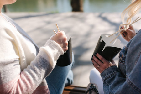 Two pretty woman eating fastfood oudtood. Beautiful girl sitting on bench in park and take asian...