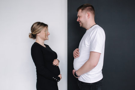 Pregnant woman compare her belly to her husbands beer belly on black and white background