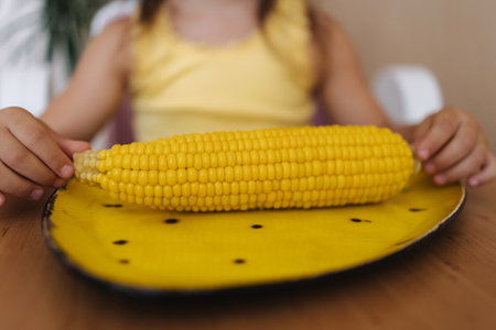 Close up of little girl hold corn in hand. Fresh corn on yellow plateの写真素材