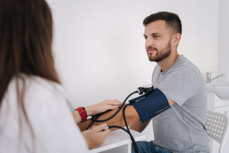 Young man with high blood pressure. Female doctor using sphygmomanometer with stethoscope checking...