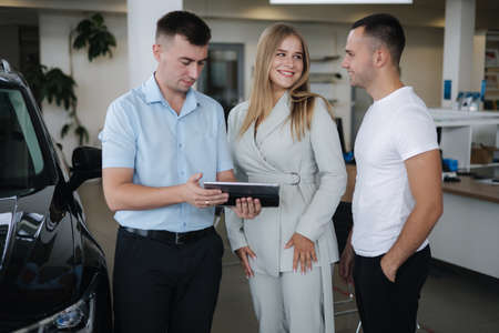 Salesperson talking with couple and help choosing new car. Man and woman in car showroom. Man using tablet to shop opportunity of different modelsの写真素材