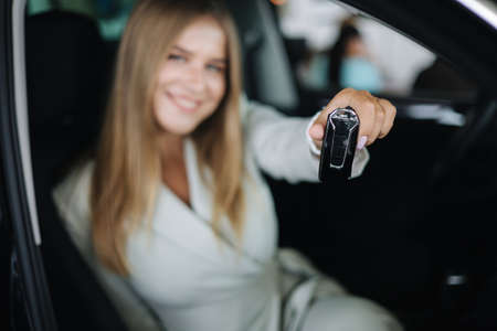 Attactive young bussines woman sitting in car in car showroom. Woman choosing new car. Beautiful blond hair female in suit hold keys. Keys in focusの写真素材