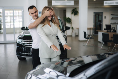 Man close eyes to his wife and makes a surprise buying car. Man and woman in car showroom