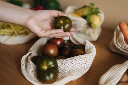 Female hand take tomato from canvas grocery bag. Vegetables in reusable eco cotton bags on wooden table. Zero waste shopping concept. Plastic free itemsの写真素材