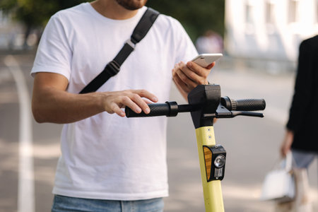 Young man connect an electric scooter outdoors. Man with black shoulder bag using phone for...