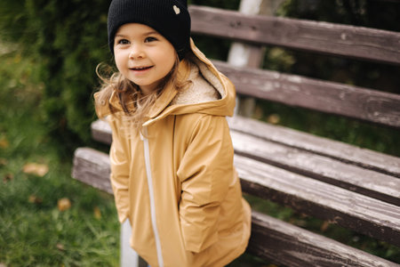 Happy cute little girl in jacket and hat posing to photographer outdoor by the bench. Little model have fun in autumn timeの写真素材