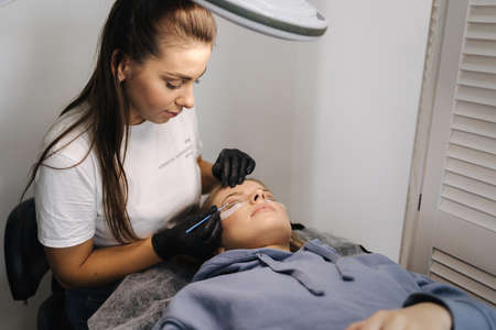 Woman doing eyelashes lamination in beauty studio. Blond hair model has staining, curling, laminating and extension for lashes. Black glovesの写真素材