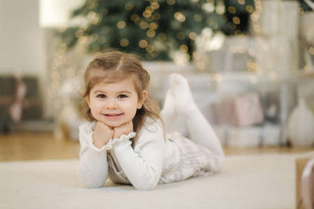 Adorable little girl lying on her front nearby fir tree and smile. Christmas mood at home with lot of presentsの写真素材