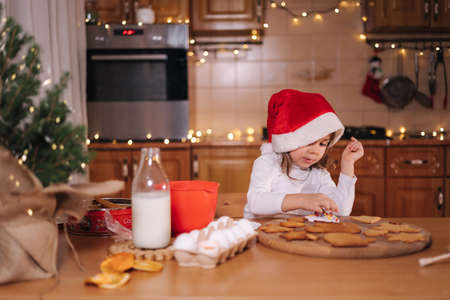 Little girl in santa hat decorates gingerbread using multicolored beads. Christmas and New Year traditions concept. Christmas bakery. Happy hollidaysの写真素材