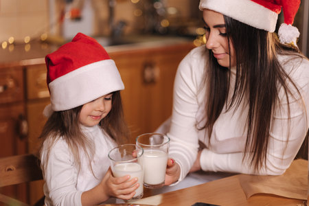 Portrait of beautiful tree year girl with her mom in santa hat sitting by the table in front of kitchen decorated fairy lights and drink fresh milk. Christmas moodの写真素材