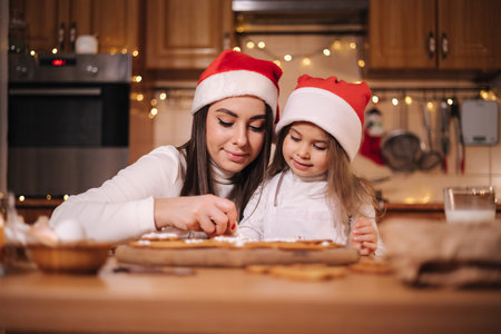 Mom in santas hat with her little doughter decorates gingerbread using glaze. Christmas and New Year traditions concept. Christmas bakery. Happy holidaysの写真素材