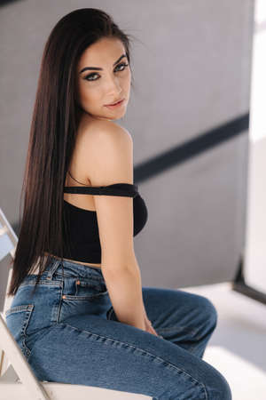 Sexy woman in black topic and blue jeans sits on chair during studio photoshoots. Brunette girl posing to photographer. Shadow on white backgroundの写真素材