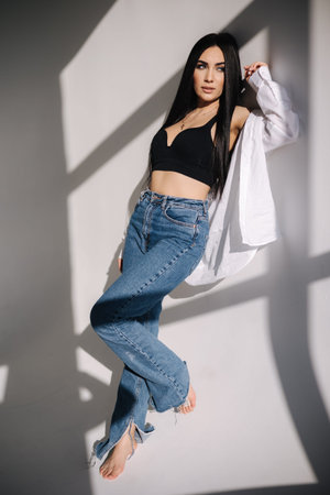 Pretty woman barefoot in studio. Beautiful brunette girl in black topic with white shirt and blue denim jeans. White background of cycloramaの写真素材