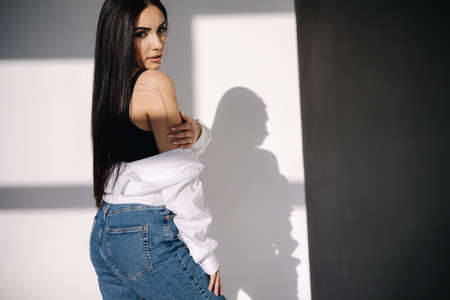 Pretty brunette hair woman in blue jeans and white shirt standing in front of white wall. Shadow on white backgroundの写真素材