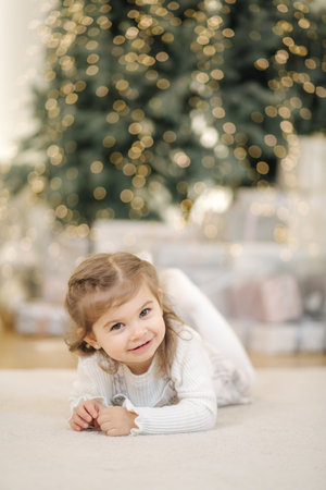 Adorable little girl lying on her front nearby fir tree and smile. Christmas mood at home with lot of presentsの写真素材
