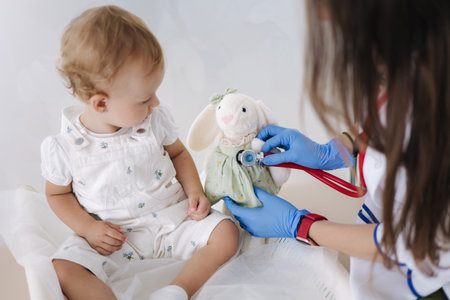Friendly female doctor checking soft toy rabbit using stethoscope. Adaptation of the little baby to the yound doctor pediatricianの写真素材