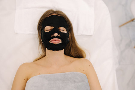 Woman with a cloth moisturizing black mask on her face in beauty studio. Face care and beauty treatmentsの写真素材