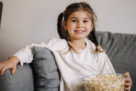 Adorable little girl watching TV at home and laughs. Cute girl eating popcorn. Holiday moodの写真素材